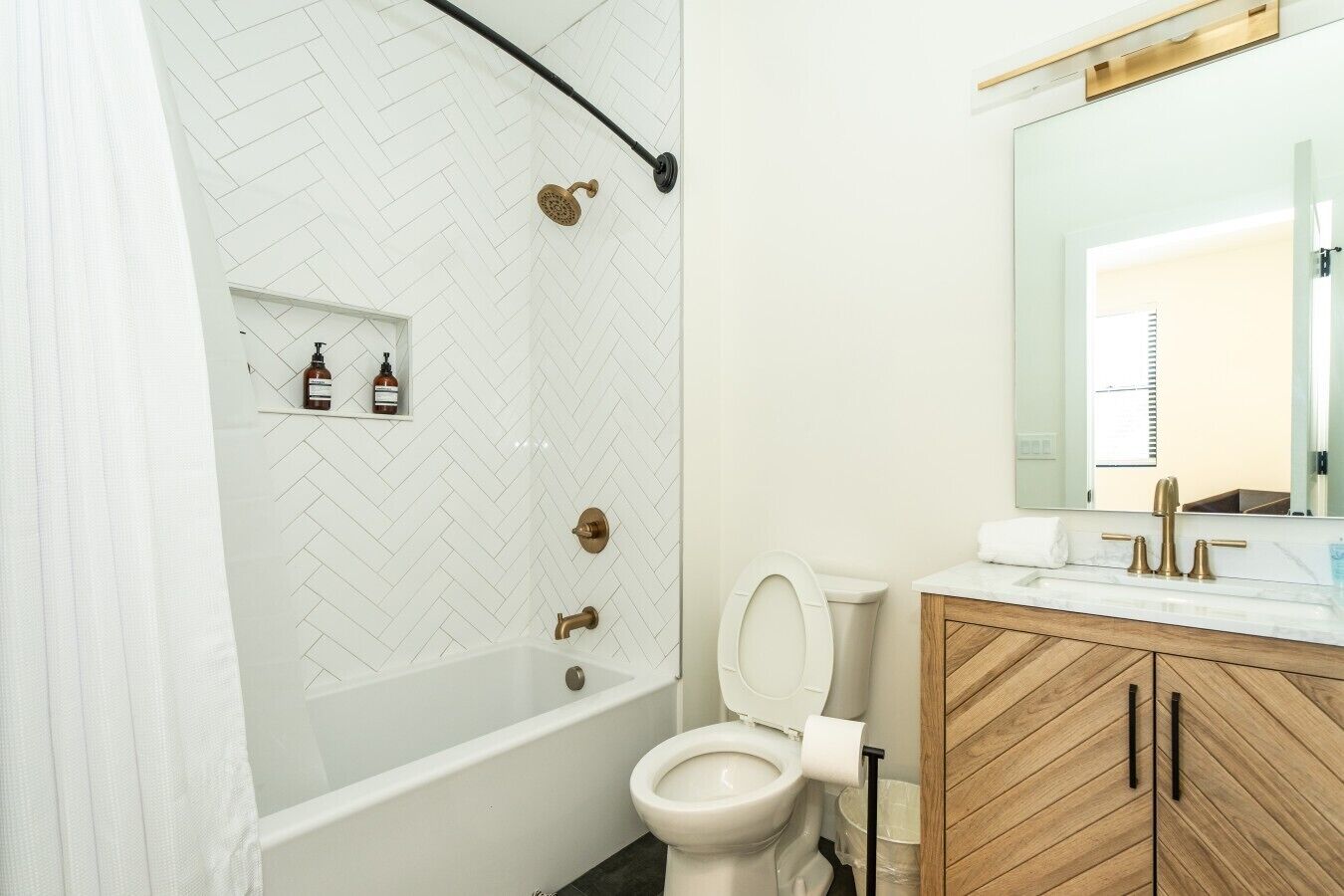 A bathroom with a white herringbone tile shower, a white toilet, a wooden vanity with a mirror, and a white bathtub.