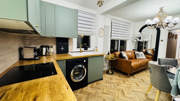 A modern kitchen with a washing machine, a wooden countertop, and a dining area with a sofa and chairs. - Designer 2 Bedroom Apartment in London with Garden (Bexleyheath)