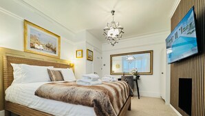 A hotel room with a large bed, a chandelier, a desk with a chair, a television, and framed artwork on the walls. - Designer 2 Bedroom Apartment in London with Garden (Bexleyheath)