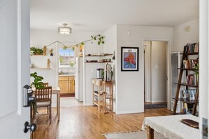 Een moderne woonkamer met houten vloer, een eethoek met tafel en stoelen, een boekenkast en een keuken die door een open deur zichtbaar is.