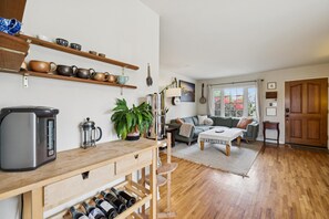 Een gezellige leefruimte met een houten salontafel, een bank, een keukenhoek met waterkoker en koffiezetapparaat, en een wijnrek.