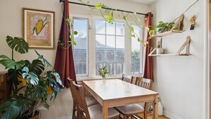 A dining area with a wooden table and chairs, a potted plant, a framed picture on the wall, and a window with curtains. - Santa Cruz Beach Bungalow | Walk to Beach | Garden (Santa Cruz)