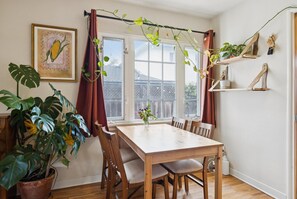 Een zithoek met een houten tafel en stoelen, een potplant, een ingelijst kunstwerk aan de muur en een raam met gordijnen.