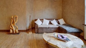 Ein Zimmer mit Holzboden, einem braunen Ledersofa mit weißen Kissen, einer Holzskulptur und einem runden Holztisch mit einem Buch und einer Fernbedienung.