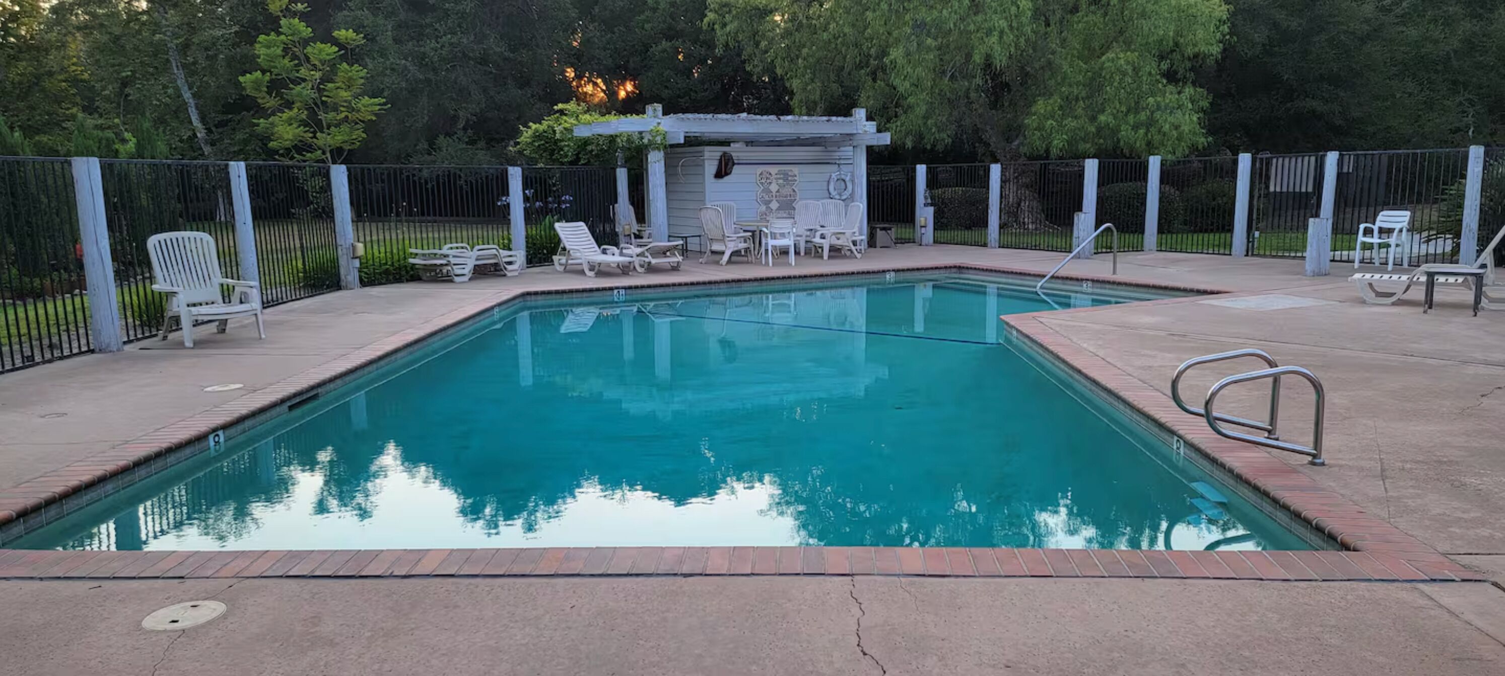 A rectangular swimming pool with a brick edge, surrounded by a metal fence.
