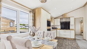 A modern kitchen with white cabinetry, a dining area with a table set for four, and a view of a wooden building outside. - Kittiwake Lodge - 2 Bed Lodge - Wisemans Bridge (Kilgetty)