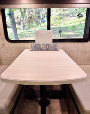 Ein Campervan-Innenraum mit einem 'WELCOME'-Schild, einem Tisch und Blick ins Freie durch die Fenster.