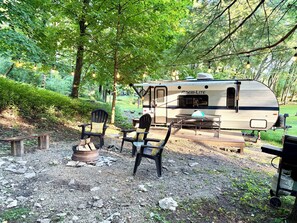 Ein Campingplatz mit Wohnmobil, zwei Adirondack-Stühlen, einer Feuerstelle und einem Picknicktisch.
