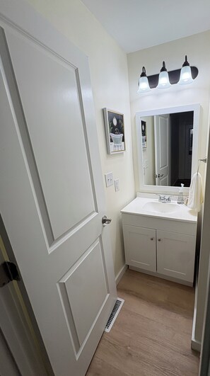 A bathroom with a white door, a sink with a mirror, and a light fixture above the mirror.