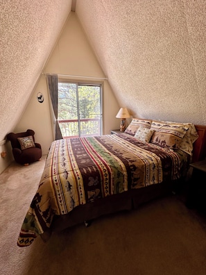 Une chambre amĂ©nagĂ©e dans les combles, avec un plafond en pente, une grande fenĂȘtre, un lit avec une literie Ă motifs, une chaise et une table de chevet avec une lampe.