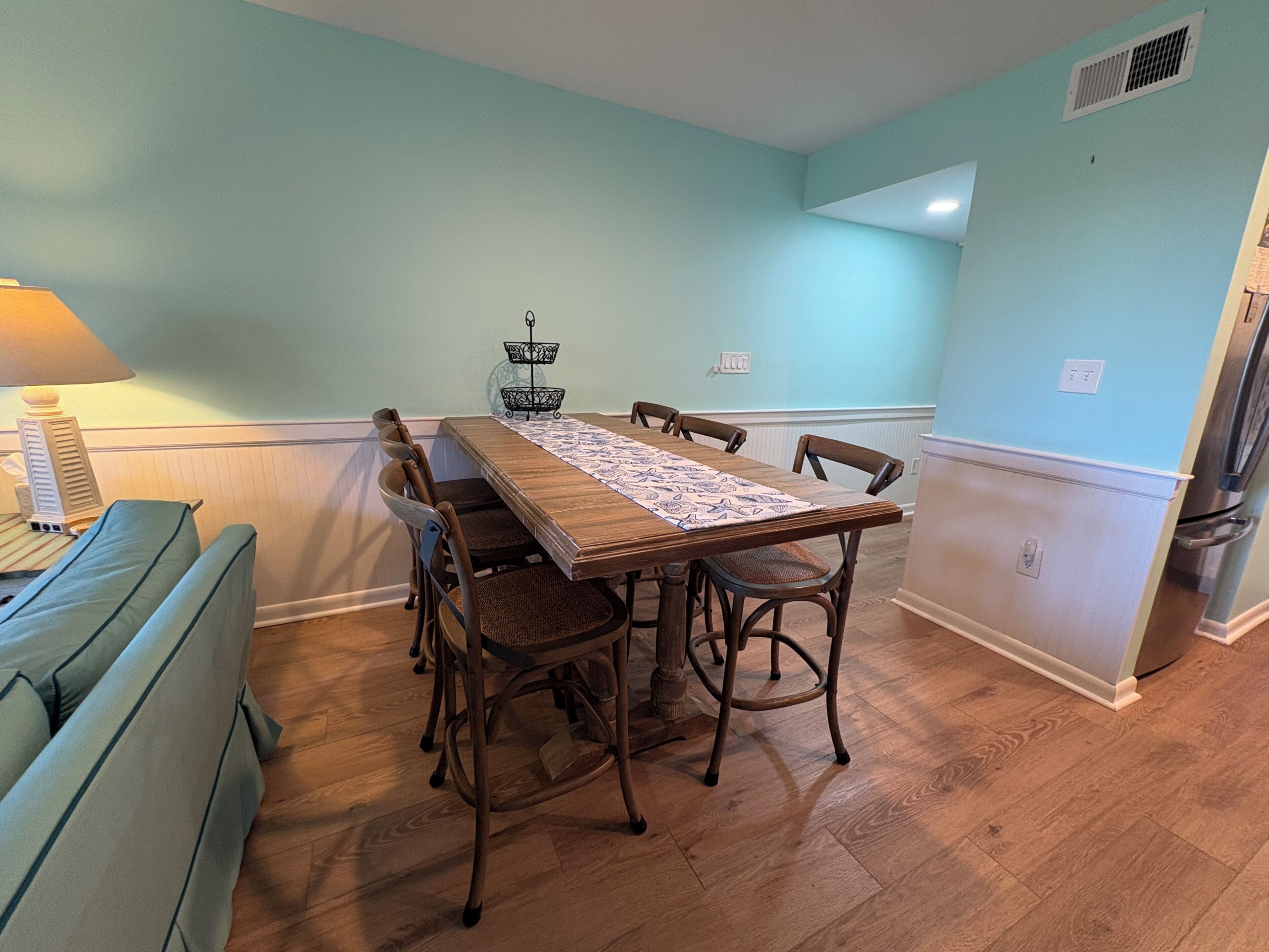A dining area with a wooden table, six chairs, a patterned runner, a small decorative basket, and a lamp.