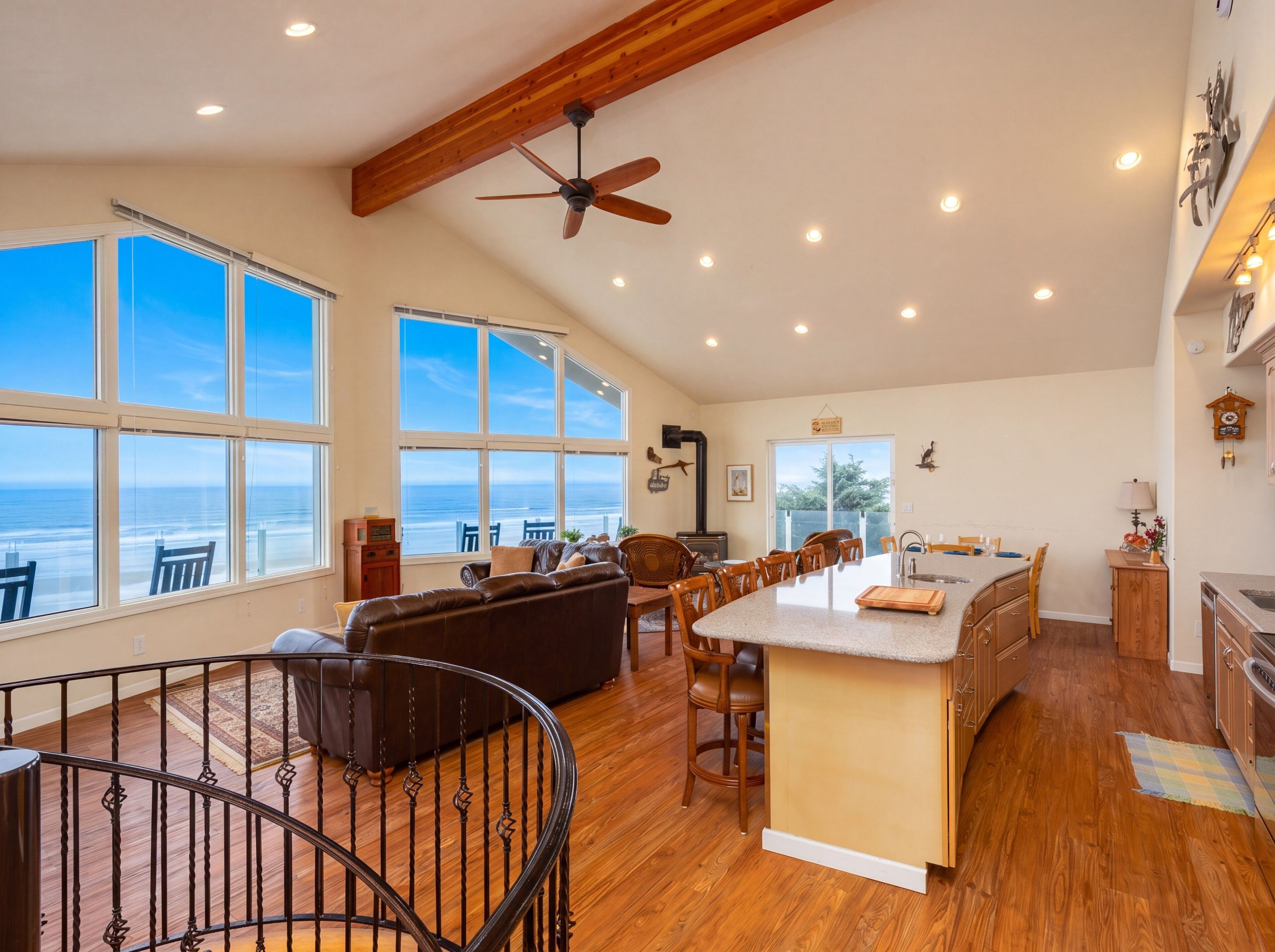 A spacious living area with a large window overlooking the ocean, a ceiling fan, and a kitchen area with wooden cabinets.