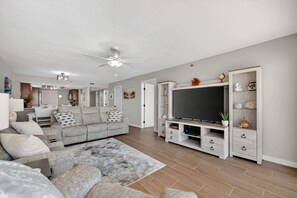 A living room with a grey sofa, a flat-screen TV, a ceiling fan, and a wooden cabinet with decorative items. - Floridian Paradise - Luxury Oceanfront Condo (Daytona Beach Shores)