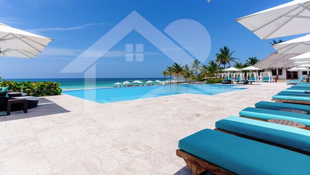 Ein Poolbereich mit Liegestühlen und Sonnenschirmen, mit Blick auf das Meer und einem Gebäude mit Reetdach im Hintergrund.