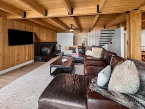 A spacious living room with a large leather sofa, a flat-screen TV mounted on the wall, and a fireplace. - Payette Lake Retreat | Dock, Beach, Room for Toys! (McCall)