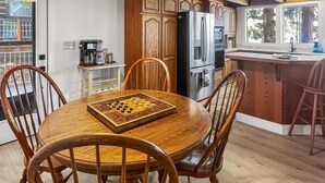 Uma mesa de jantar de madeira com cadeiras, uma cafeteira e um jogo de xadrez em uma cozinha com armários de madeira e uma geladeira.