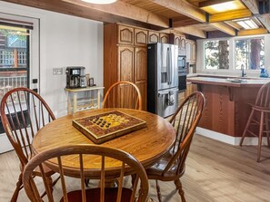 A wooden dining table with chairs, a coffee maker, and a chessboard in a kitchen with wooden cabinets and a refrigerator. - Payette Lake Retreat | Dock, Beach, Room for Toys! (McCall)