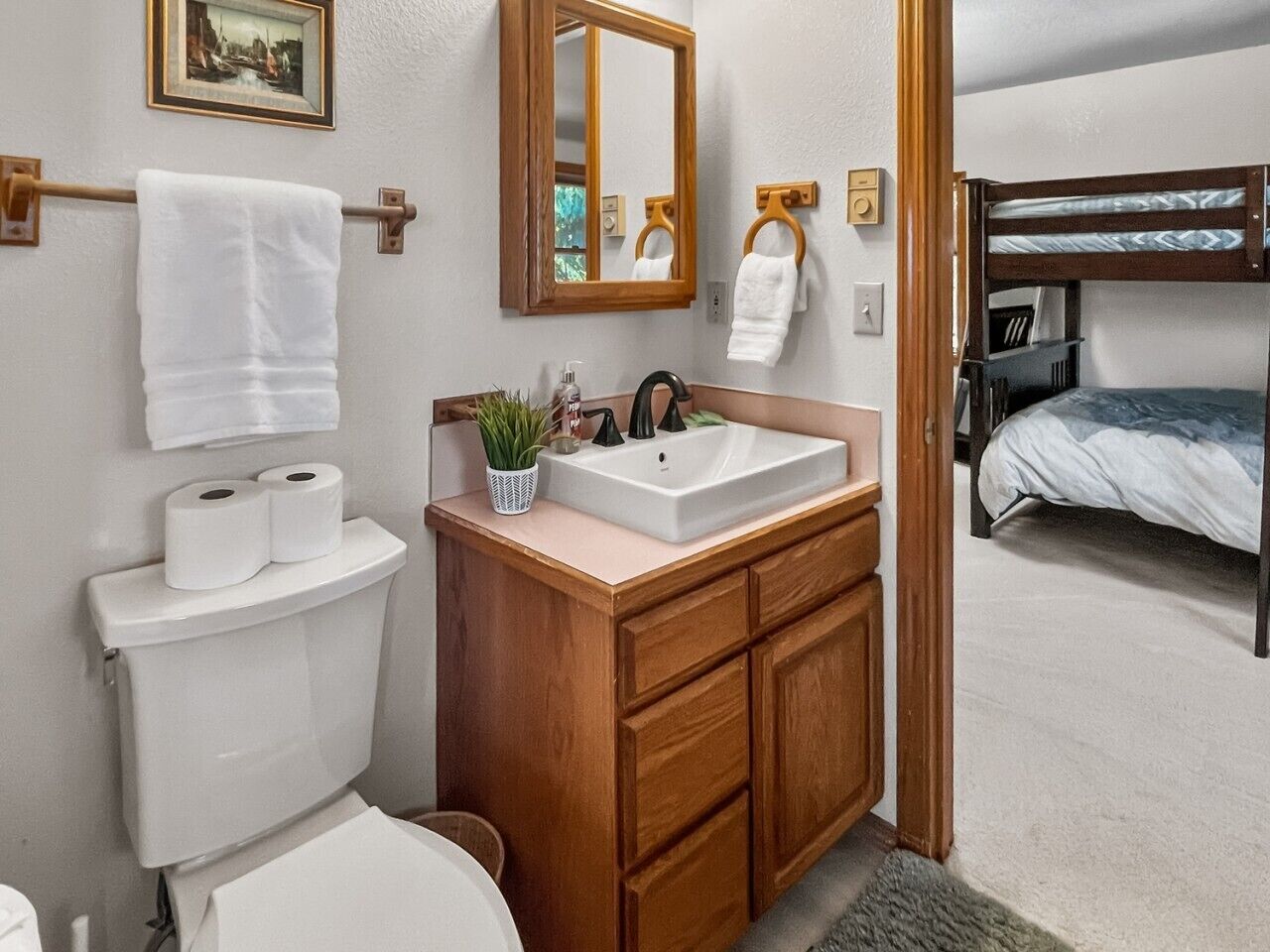 Casa de banho com sanita, lavatório e espelho. Existe uma cama beliche na sala adjacente.
