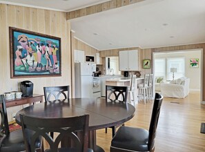 Ein Essbereich mit rundem Tisch und Stühlen, eine Küche mit weißen Schränken sowie ein Wohnzimmer mit weißem Sofa und einem Gemälde.
