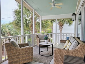 Eine überdachte Veranda mit Korbmöbeln, einem Deckenventilator und Blick auf Palmen.