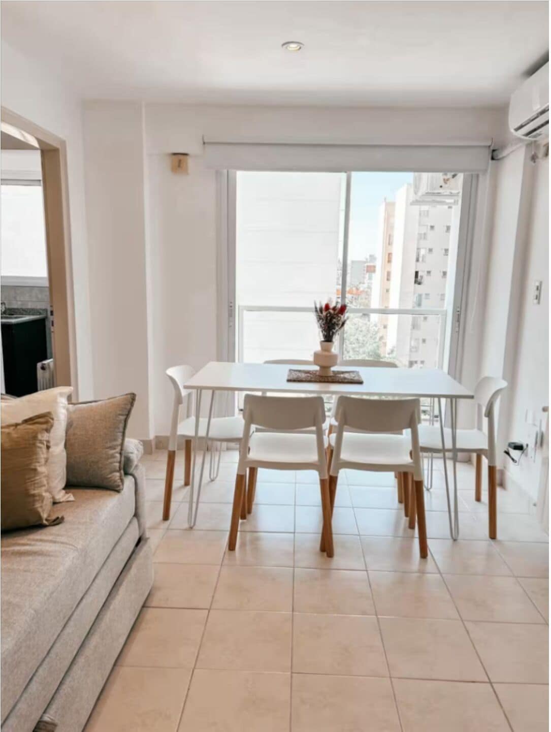 A modern living room with a dining table, white chairs, a sofa, and a large window with a city view.