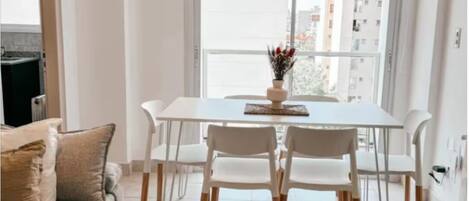 A modern living room with a dining table, white chairs, a sofa, and a large window with a city view.