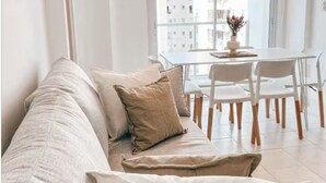 A modern living room with a sofa, a dining table with chairs, and a view of buildings through large windows. - Dpto ubicación ideal para 6 (Salta)