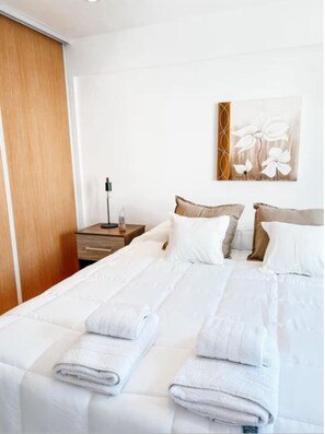 A neatly made bed with white linens and towels, a wooden nightstand with a lamp, and a wall art piece. - Dpto ubicación ideal para 6 (Salta)