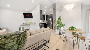 Un salon moderne avec un canapé, un téléviseur, un coin repas comprenant des chaises et une table, et une cuisine en arrière-plan.