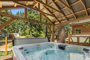 Ein Whirlpool unter einer Holzpergola mit Blick auf eine Hütte und einen Wald.