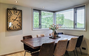 A dining room with a wooden table, six chairs, a large wall clock, and a window with blinds.