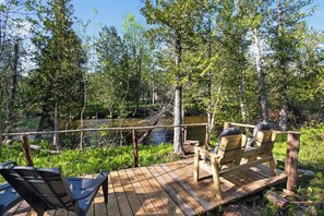 Eine Holzterrasse mit zwei Liegestühlen, einem Picknicktisch und Blick auf eine bewaldete Landschaft mit einem Bach.