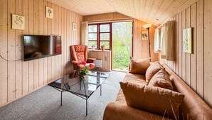 Una habitación con paneles de madera, un sofá, una mesa de centro de vidrio, un sillón de cuero, un televisor y una ventana con vistas a la vegetación.