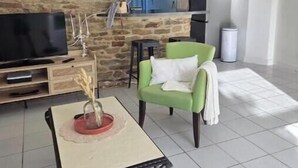 Sala de estar com parede de pedra, televisão, uma poltrona verde, uma mesa pequena e cozinha ao fundo.