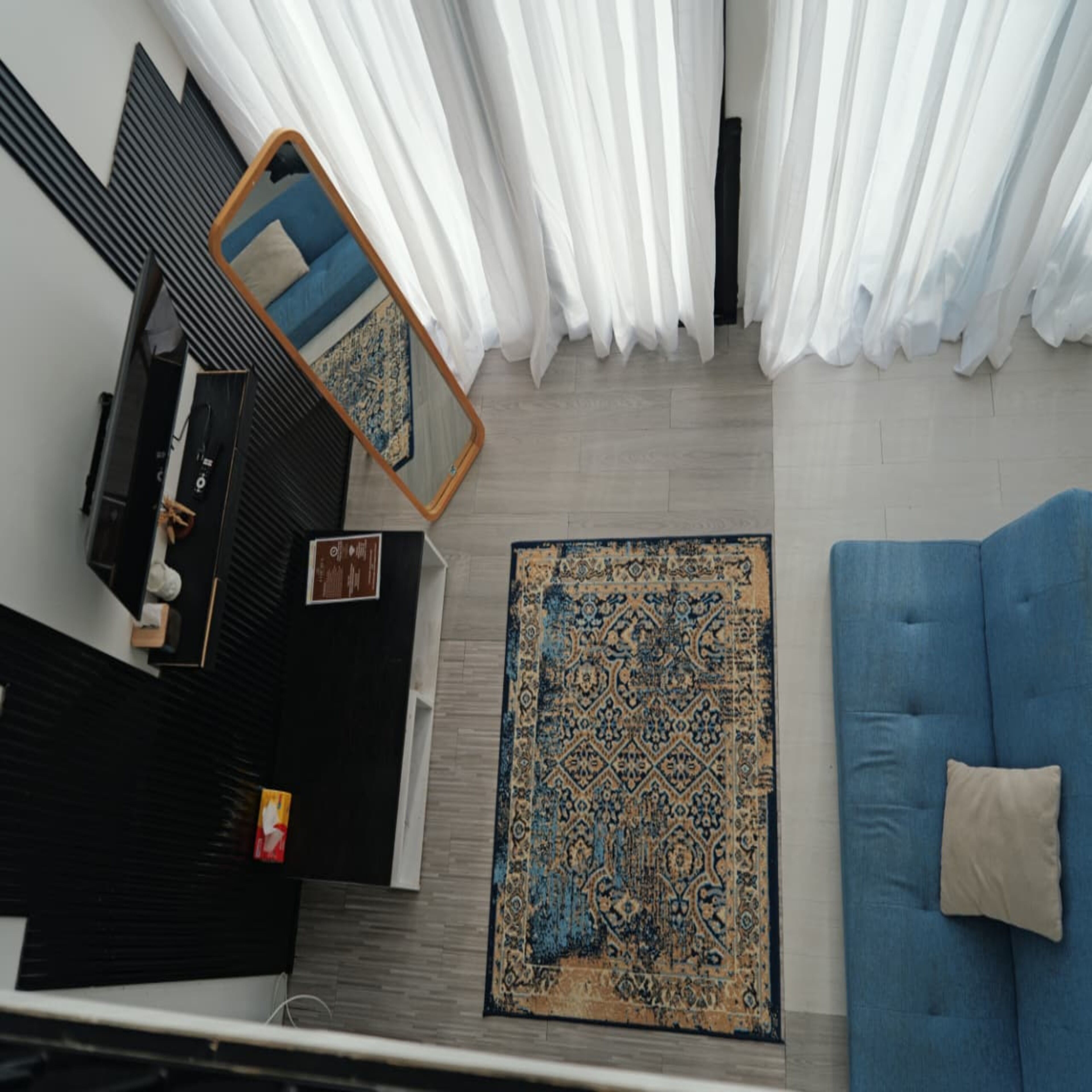 A room with a blue sofa, a patterned rug, a mirror, and white curtains.