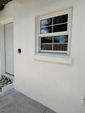 Pared exterior blanca con una ventana, una puerta y un escalón pequeño que lleva a la entrada con baldosas.
