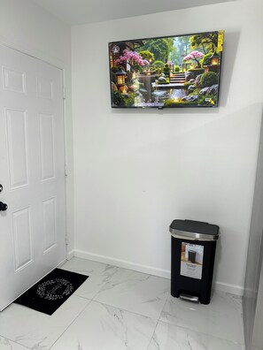 A room with a white door, a black trash bin, a black doormat, and a TV mounted on the wall displaying a garden scene.