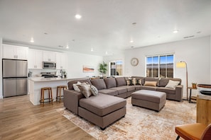 A modern living room with a sectional sofa, a kitchen area with white cabinets and stainless steel appliances, and a large window with a view. - Brand New Skyline Resort 1737, Private Hot tub, Resort Access, & Near Sand Hollow Lake (Washington)