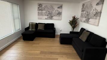 Een ruime zithoek met een zwarte modulaire bank, een salontafel en een potplant. Twee grote wanddecoraties en een raam met zonwering.