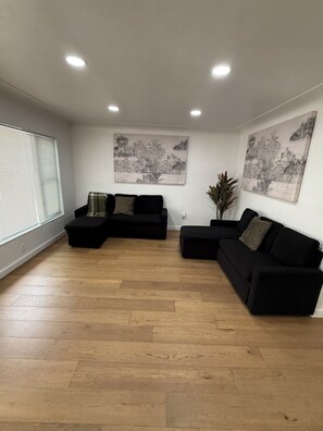 A spacious living area with a black sectional sofa, a coffee table, and a potted plant. There are two large wall-mounted paintings and a window with blinds.