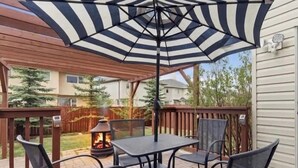 A patio with a table and chairs, a fire pit, and a large striped umbrella. - King Bed, 75 min Banff, Backyard and Wood Fire BBQ (Calgary)