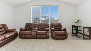 A room with a high ceiling, a large window with blinds, a brown leather sofa, a small table, and a chair. - King Bed, 75 min Banff, Backyard and Wood Fire BBQ (Calgary)