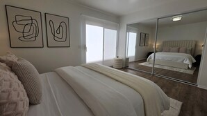 Ett sovrum med en stor spegel, en säng med vita sängkläder, en rund pall och abstrakt väggkonst.