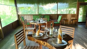 A wooden dining table set with plates, glasses, and a bottle, surrounded by chairs in a covered outdoor area with a view of greenery.