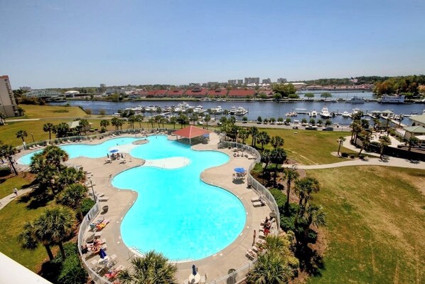 A large outdoor swimming pool with multiple sections, surrounded by palm trees and lounge chairs. - Golf course view condo with screened-in patio, W/D, pool & close to beach (North Myrtle Beach)