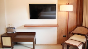 Ein Zimmer mit Flachbildfernseher an der Wand, einer Holzbank, einem Korbstuhl, einem kleinen Tisch mit einem gewebten Korb und einer Stehlampe.