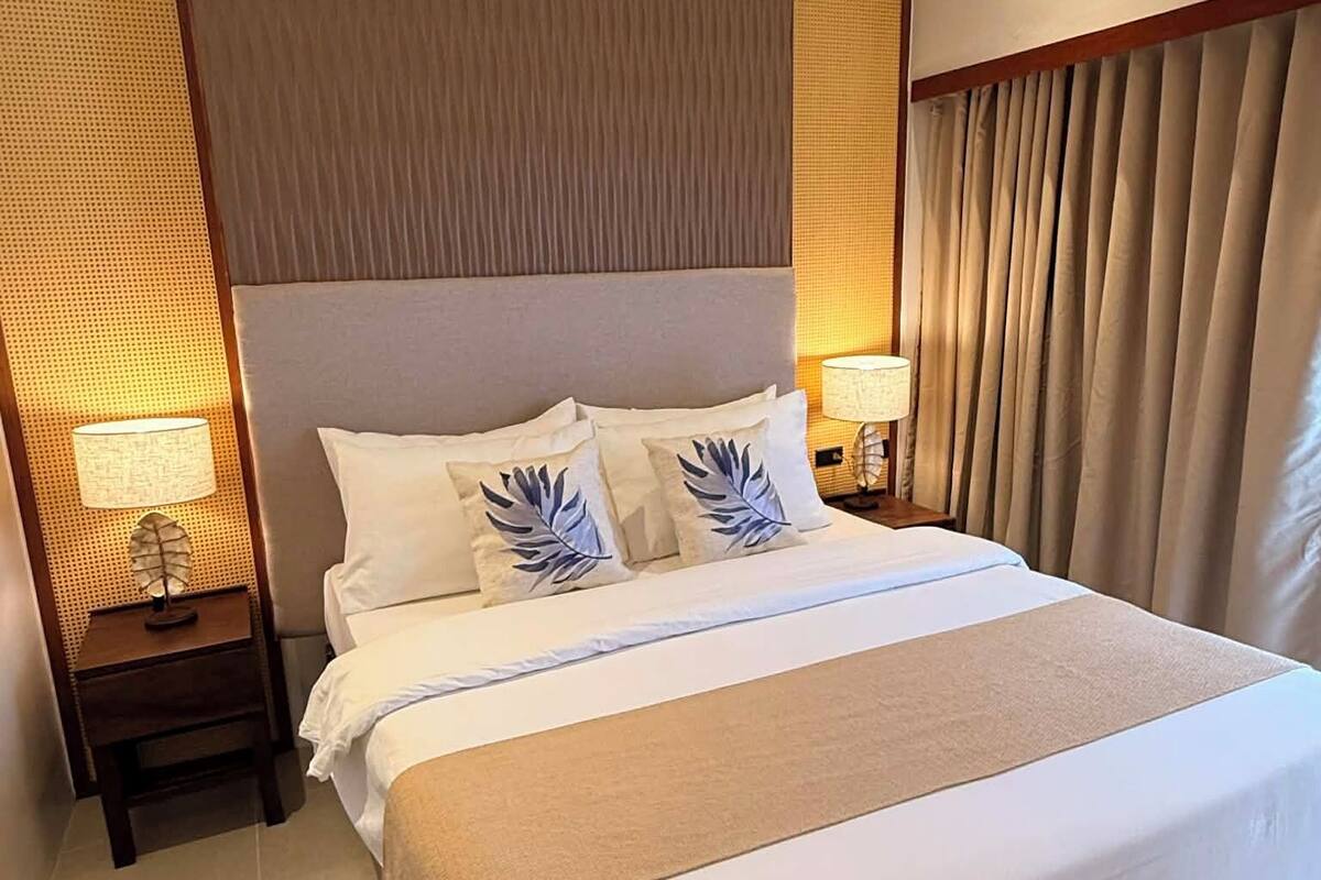 Habitación de hotel con una cama grande, una mesita de noche con lámpara y vistas al exterior a través de cortinas.