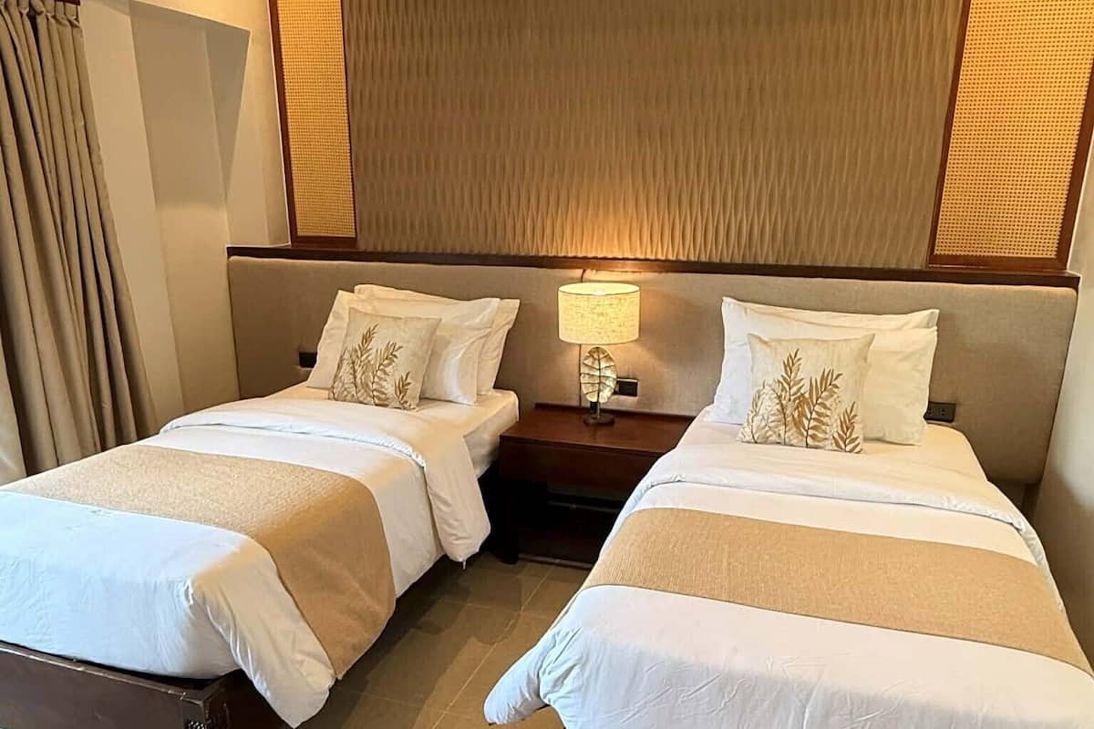 Una habitación de hotel con dos camas, una mesita de noche con lámpara y un ventanal con cortinas.