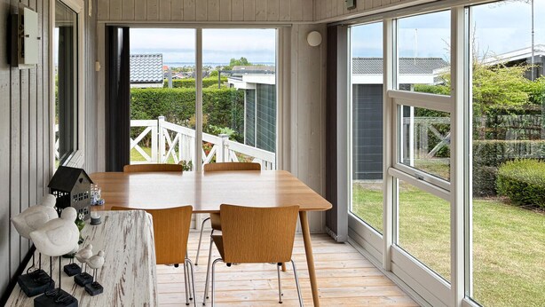 Un espace repas avec une table et des chaises en bois, de grandes fenêtres et une vue sur un jardin et des bâtiments.