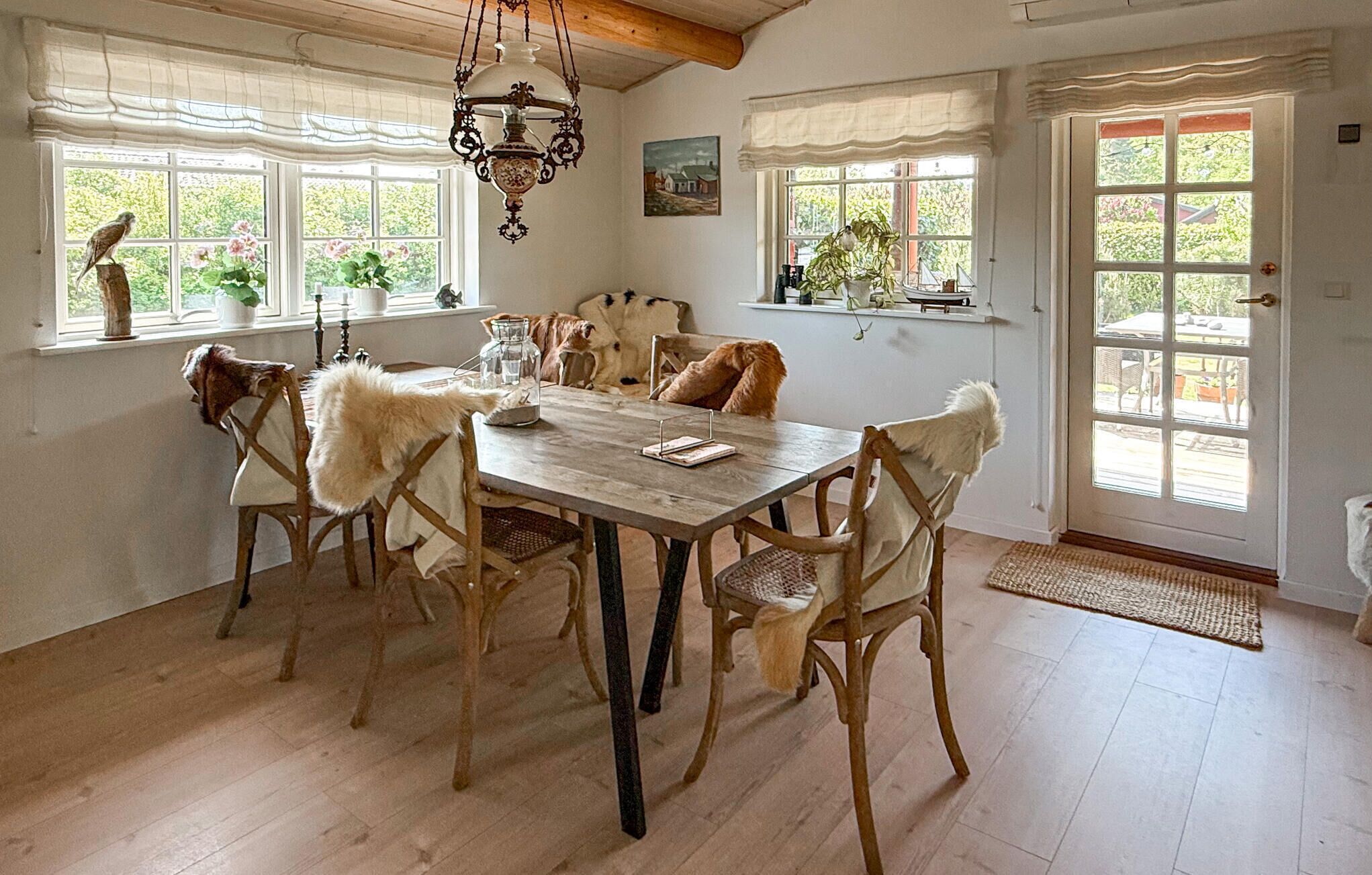 A dining room with a wooden table and chairs, a hanging light fixture, and windows with curtains.